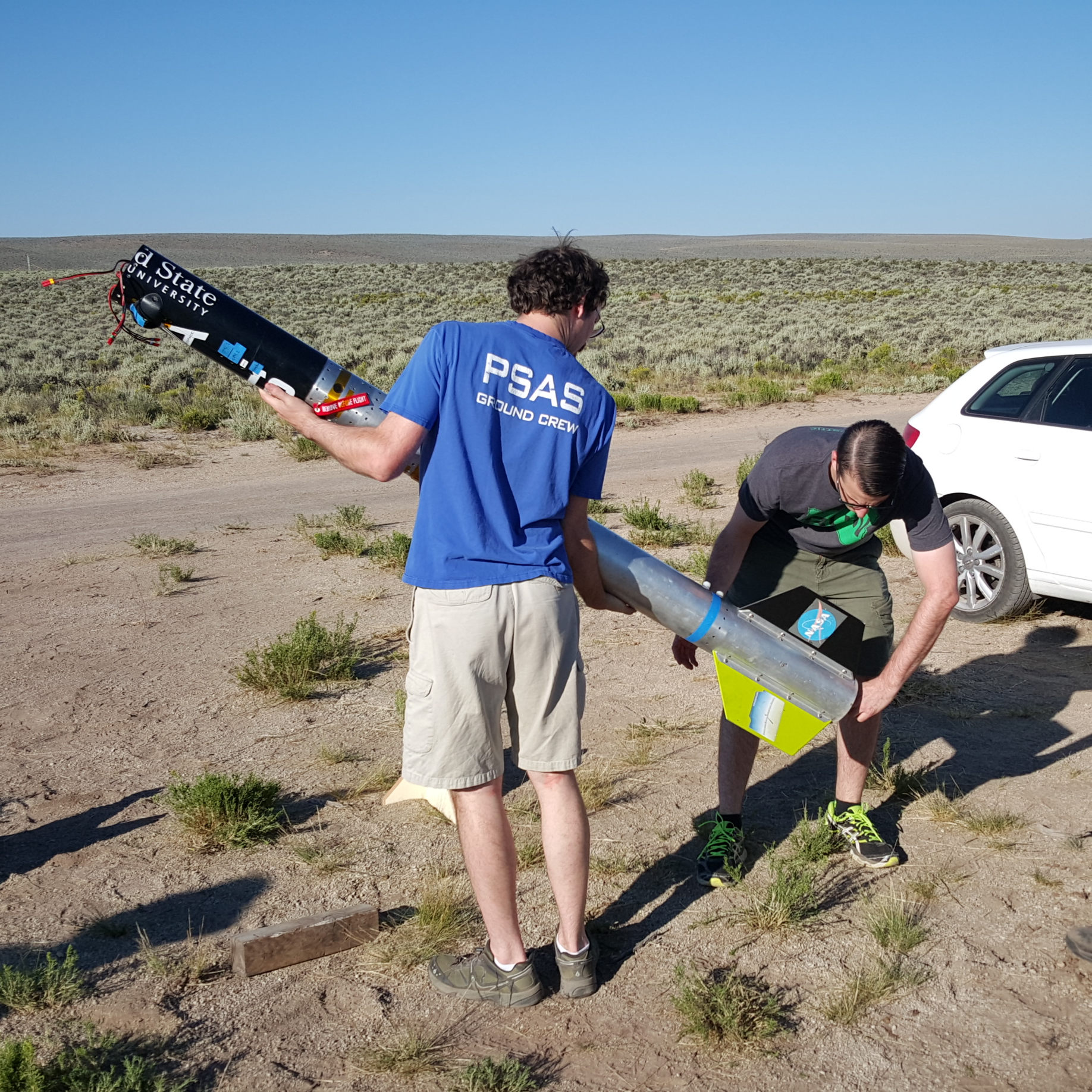 photo of two men awkwardly holding a large rocket body and an angle